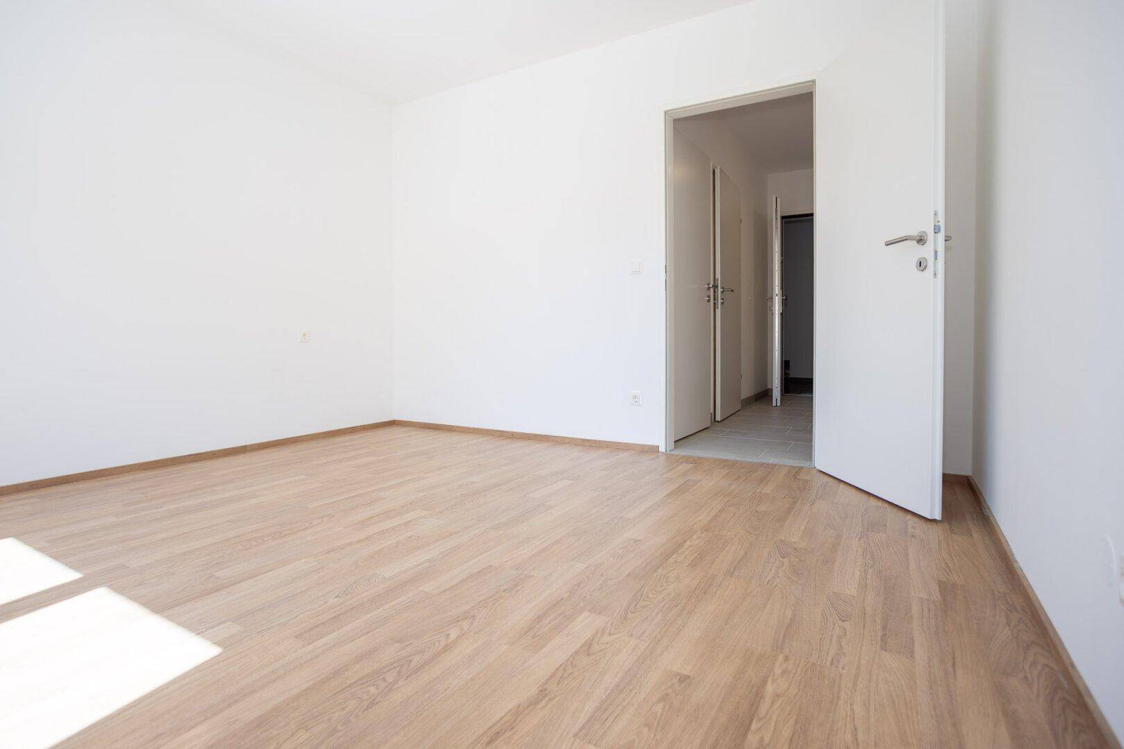 Unmöbliertes Schlafzimmer mit Laminatboden und weiß gestrichenen Wänden, mit Zugang zum Flur.