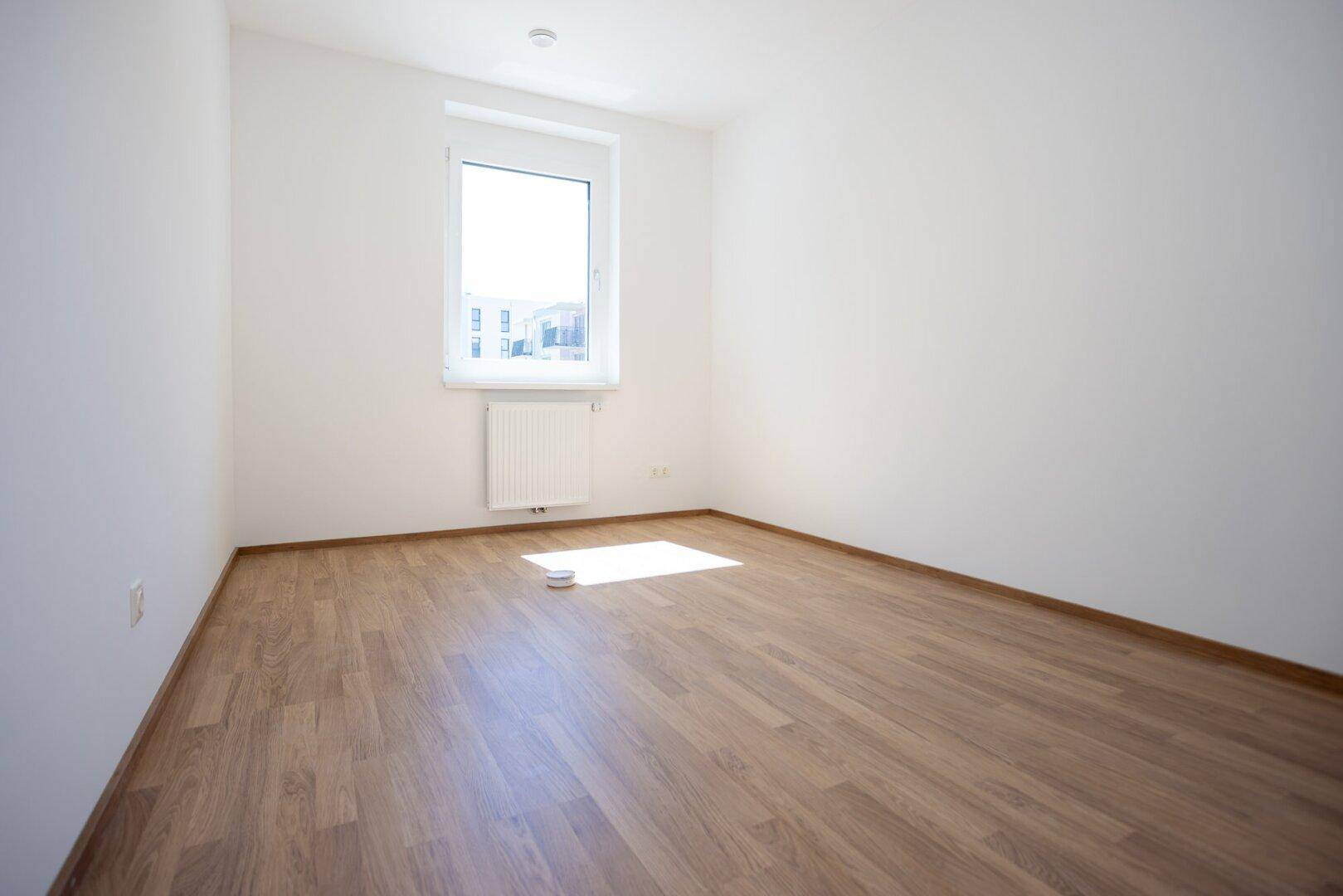 Leeres Schlafzimmer mit hellem Laminatboden und einem Fenster, das für natürliche Beleuchtung sorgt.