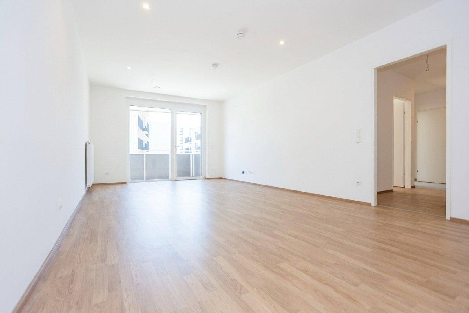 Helles, leeres Wohnzimmer mit hellem Laminatboden und Zugang zu einem Balkon.