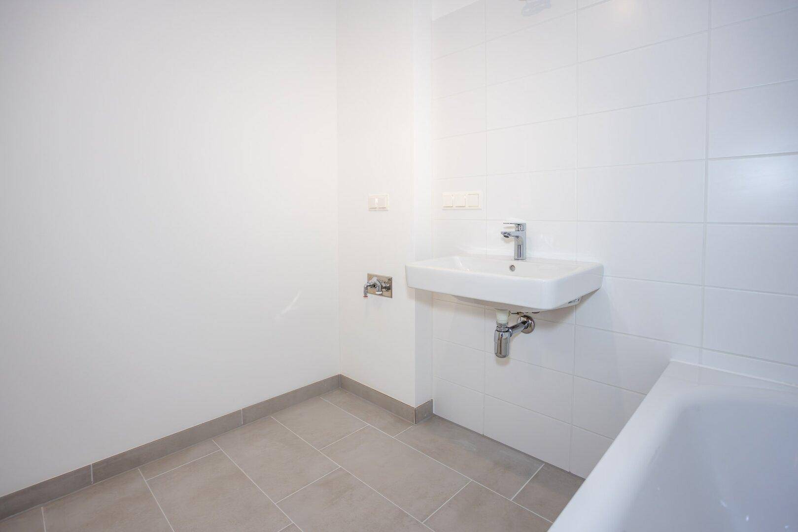 Helles Badezimmer mit Waschbecken, weißen Wandfliesen und grauen Bodenfliesen im Neubau.