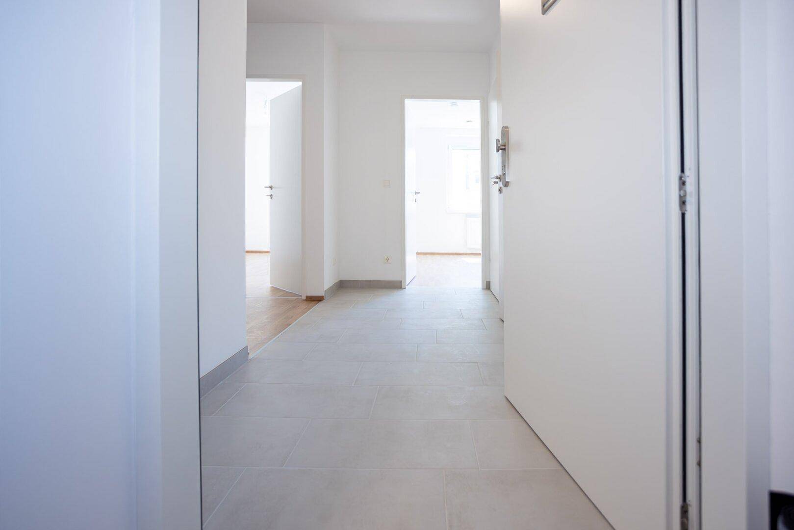 Heller Flur mit grauen Fliesen und offenen Türen zu verschiedenen Räumen im Neubau.