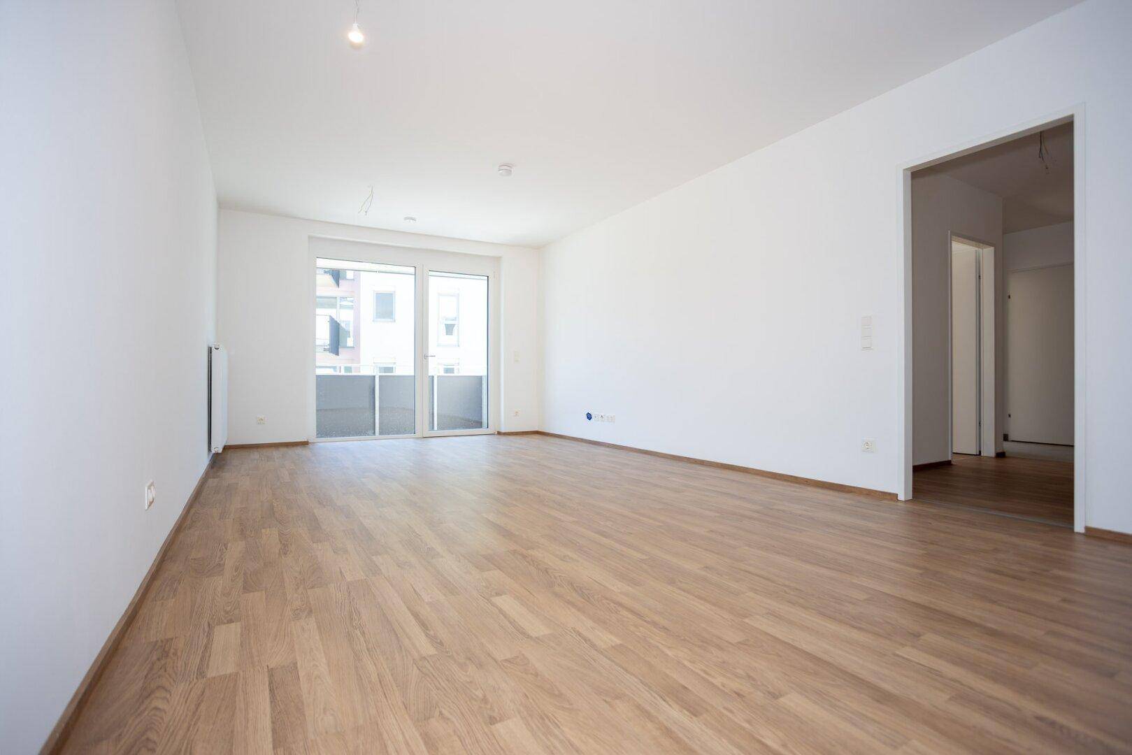 Leeres Wohnzimmer mit Laminatboden, weiß gestrichenen Wänden und Zugang zu weiteren Räumen.