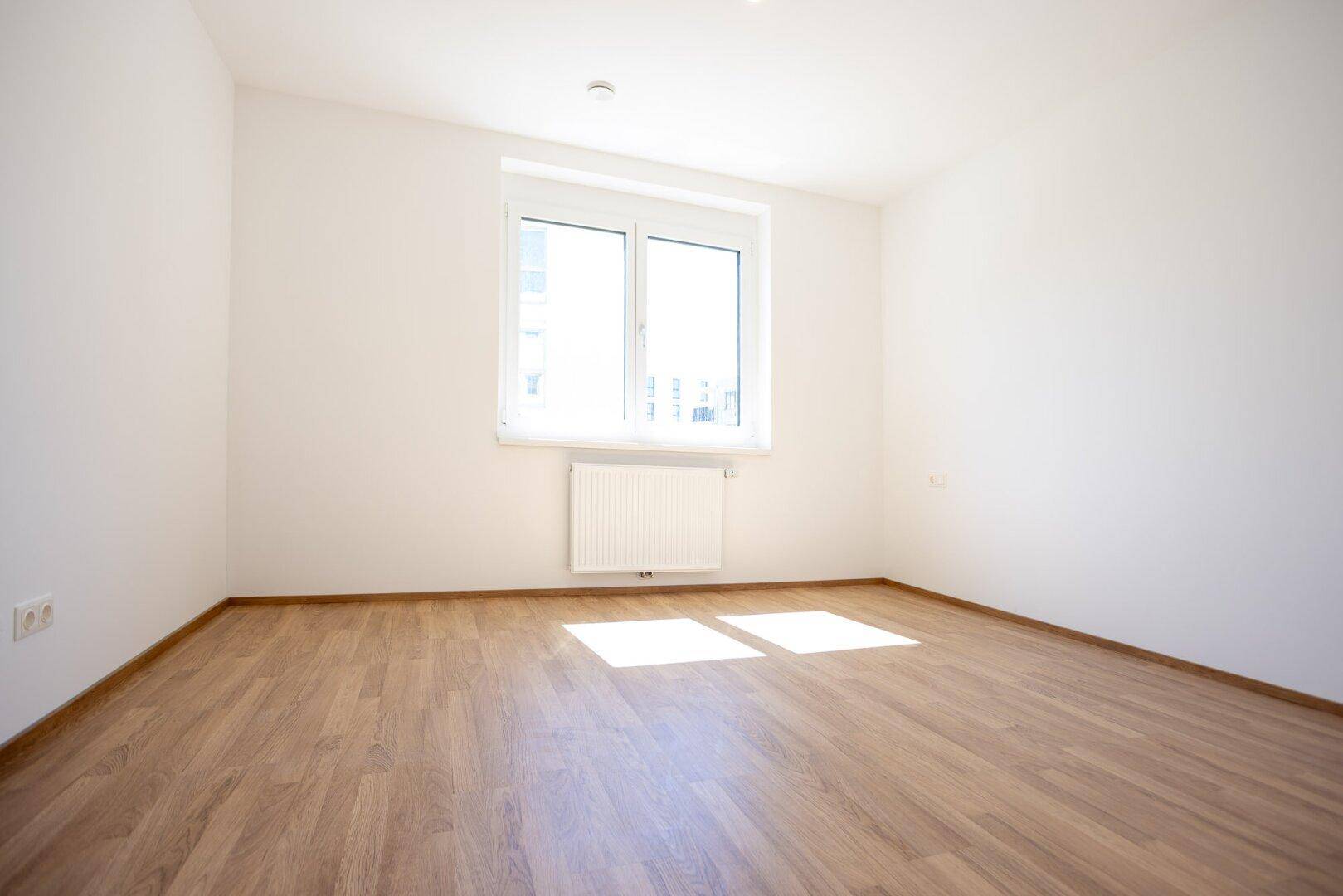 Helles Schlafzimmer mit Laminatboden, weiß gestrichenen Wänden und großem Fenster für Tageslicht.