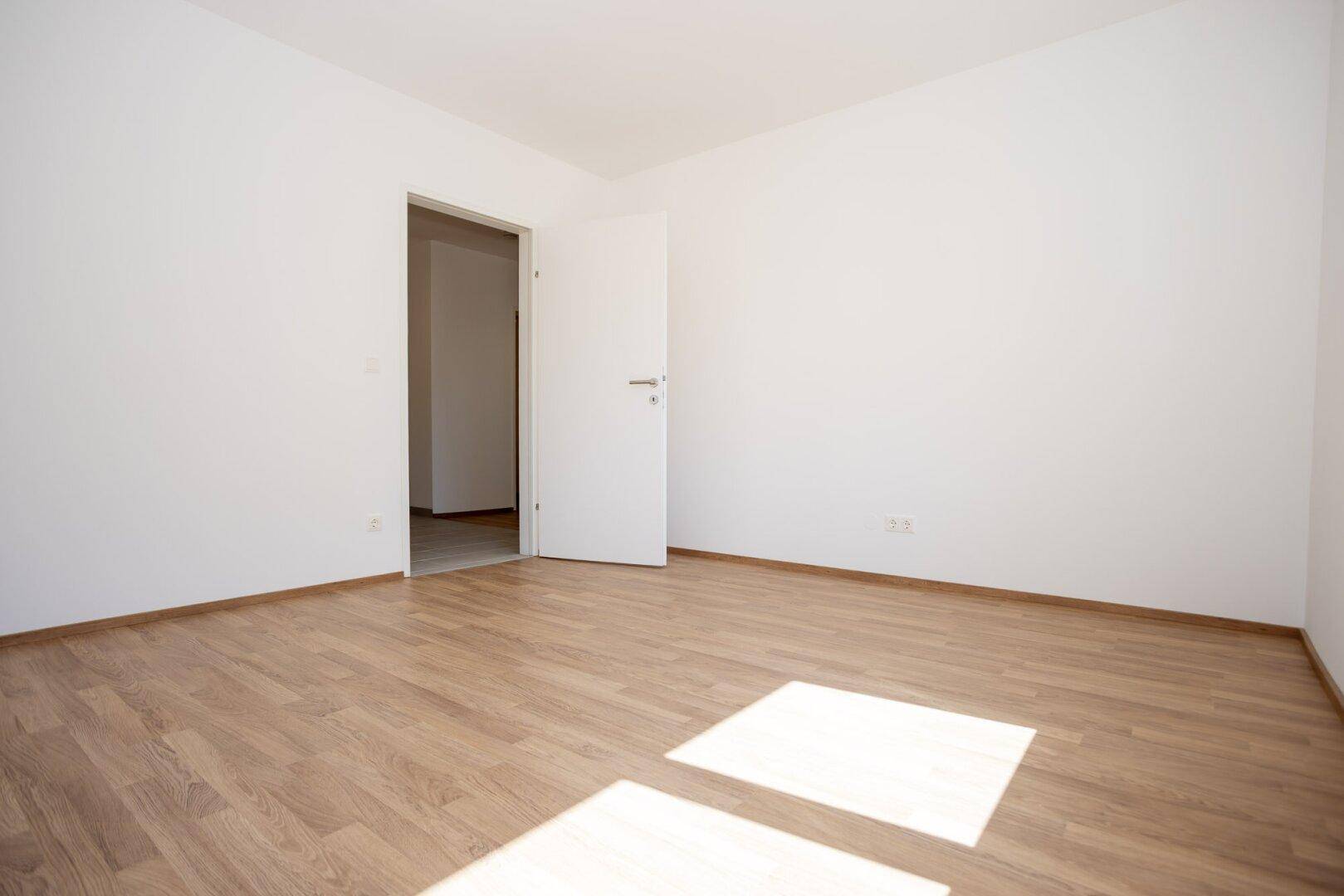 Leeres Schlafzimmer mit Laminatboden und offener Tür zu einem angrenzenden Raum im Neubau.