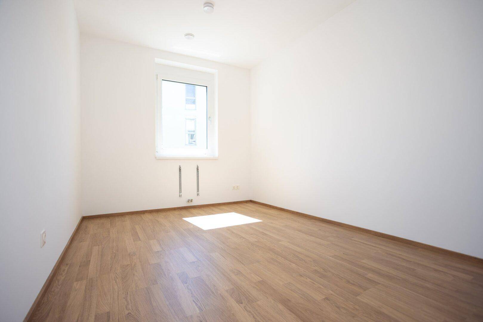 Helles Schlafzimmer mit Laminatboden und einem großen Fenster, das viel Licht spendet.