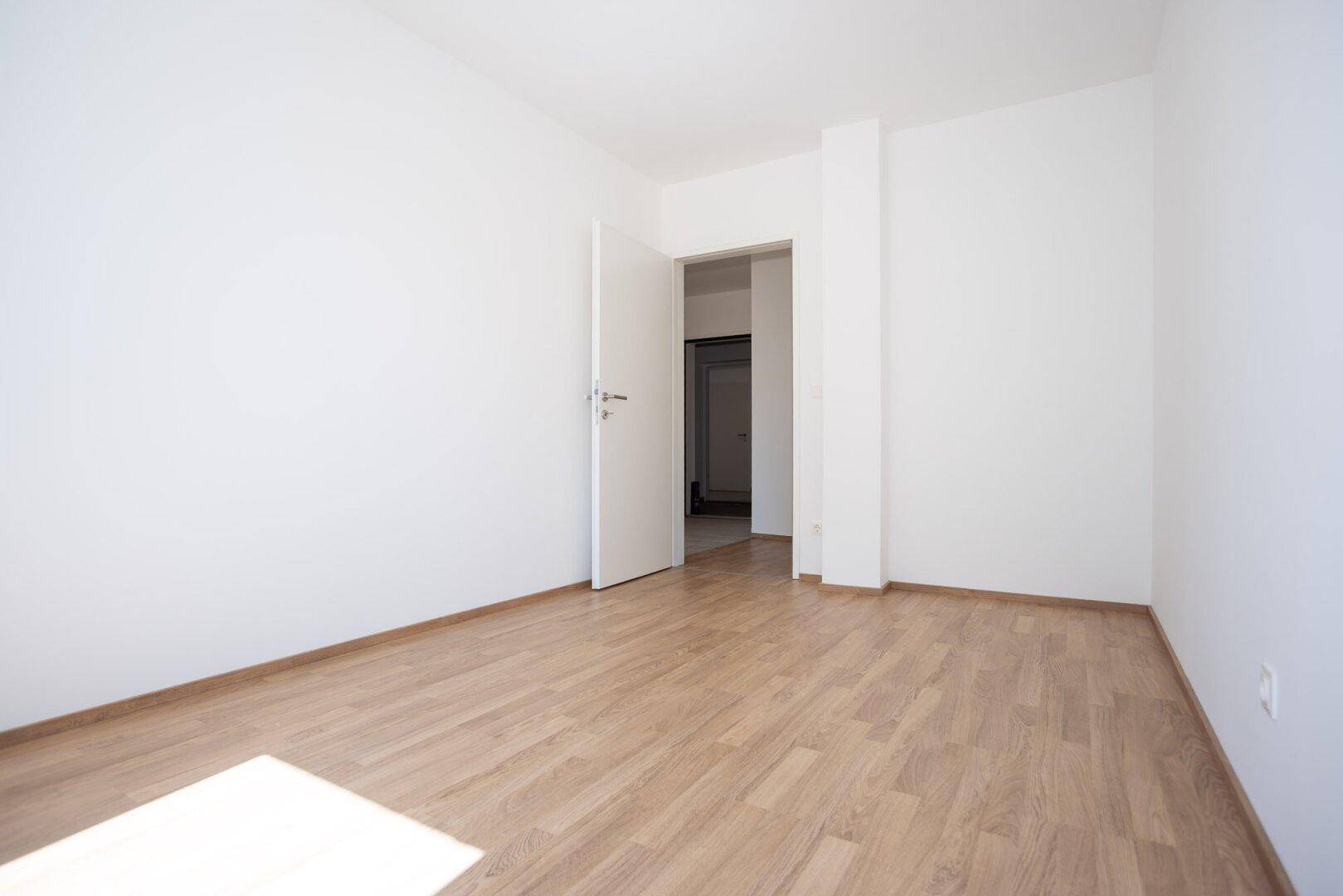 Leeres Schlafzimmer mit Laminatboden und offener Tür zu einem weiteren Raum im Neubau.