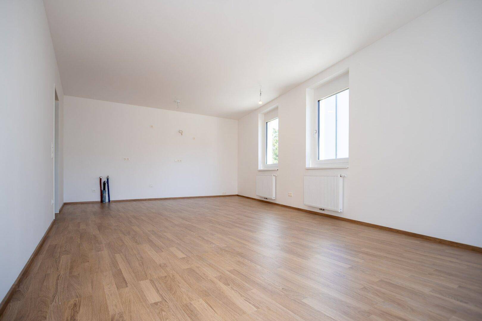 Helles, leeres Zimmer mit zwei Fenstern, Heizkörpern und Laminatboden in einem Neubau.