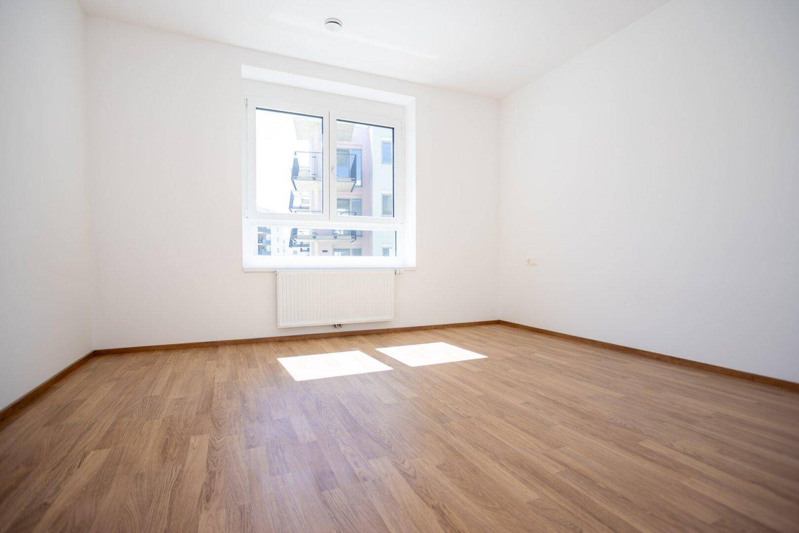 Helles Schlafzimmer mit Laminatboden, weiß gestrichenen Wänden und großem Fenster.