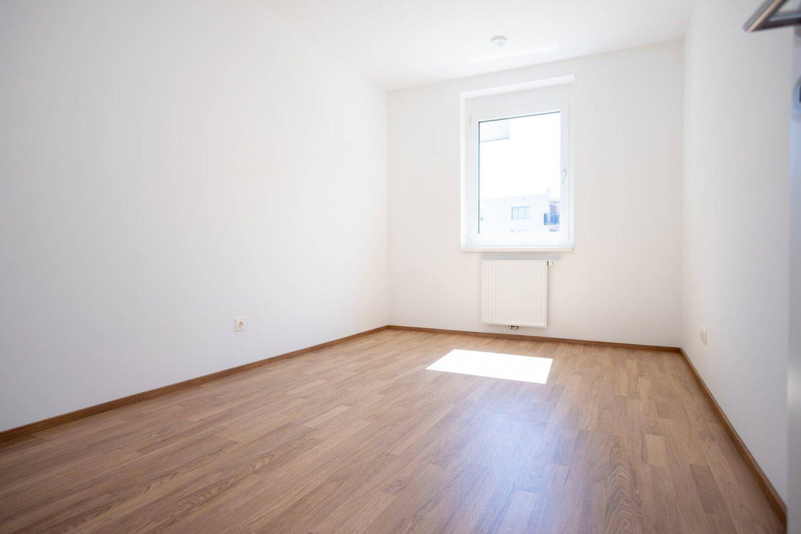 Helles Schlafzimmer mit Laminatboden, weiß gestrichenen Wänden und einem Fenster.