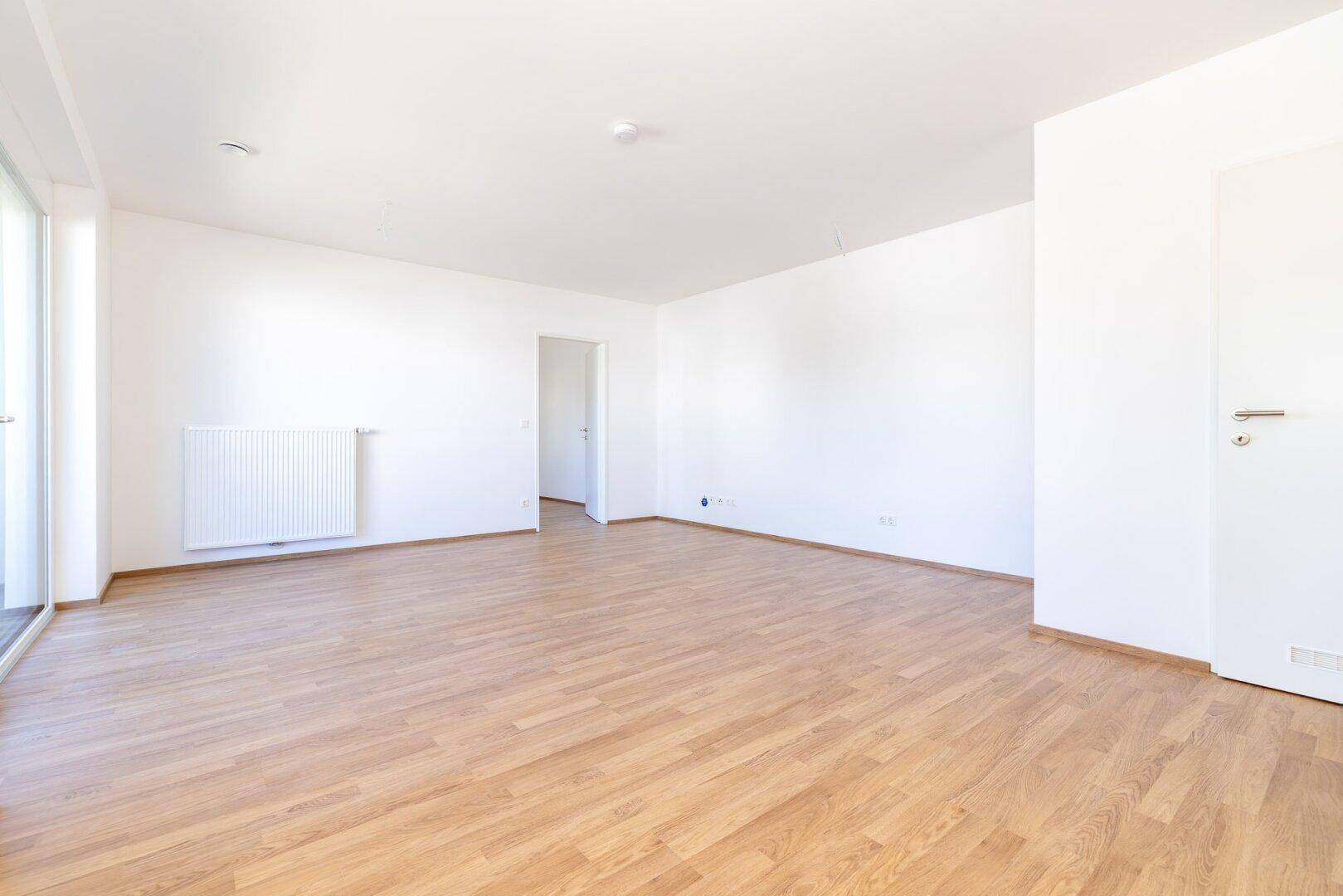 Leerer Wohnraum mit Laminatboden, weiß gestrichenen Wänden und einem Heizkörper.