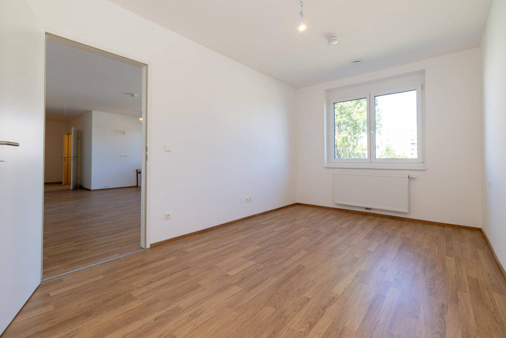 Helles Schlafzimmer mit Laminatboden und einem Fenster mit Blick ins Grüne.