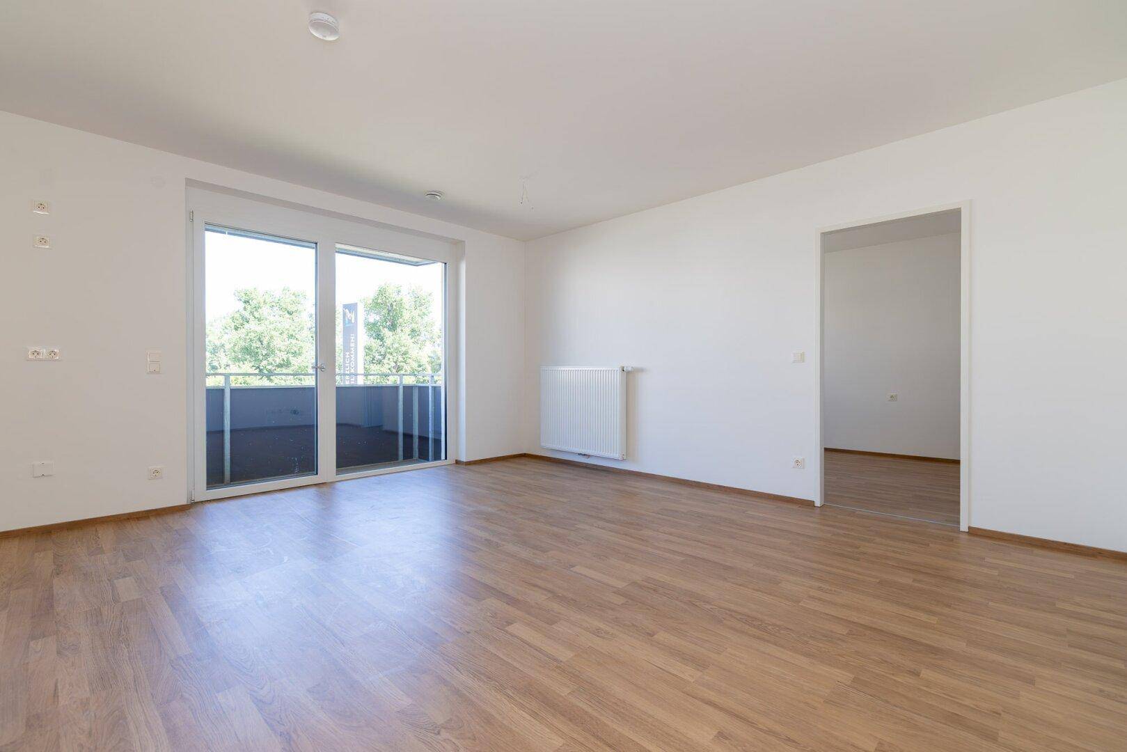 Geräumiges, leeres Wohnzimmer mit hellem Laminatboden und großem Zugang zum Balkon.