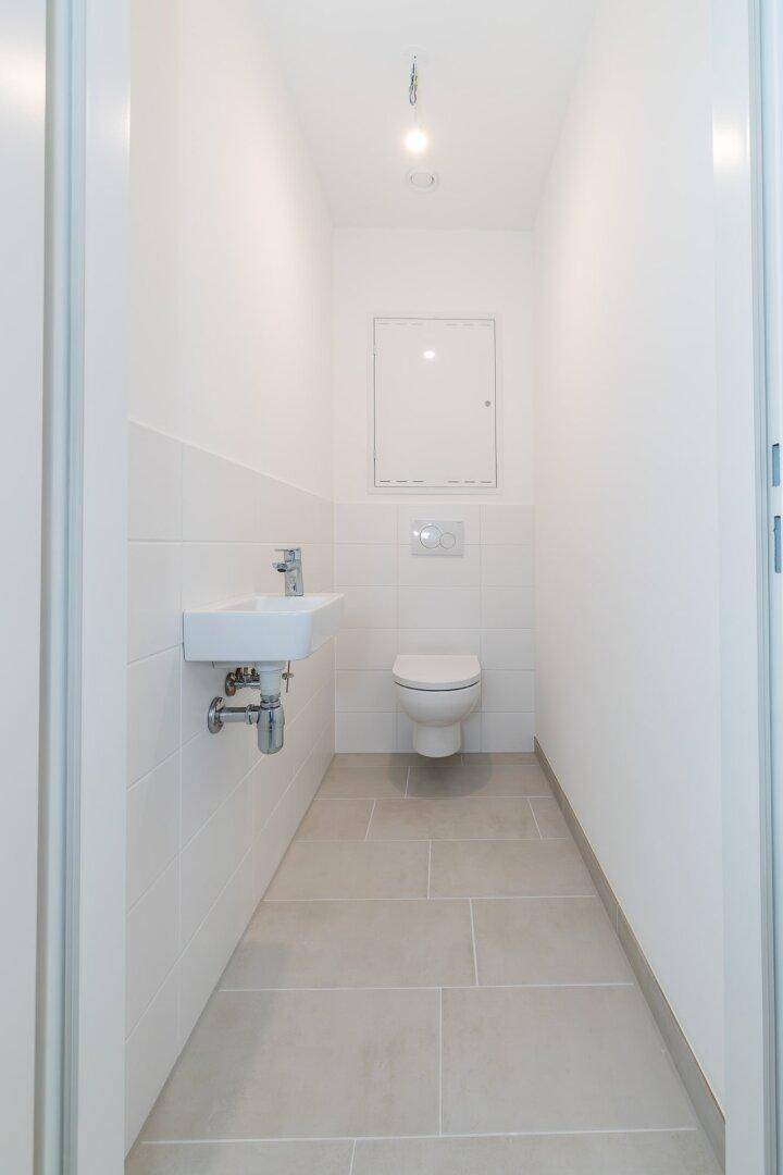 Separates WC mit wandhängender Toilette, kleinem Waschbecken und Fliesenboden.
