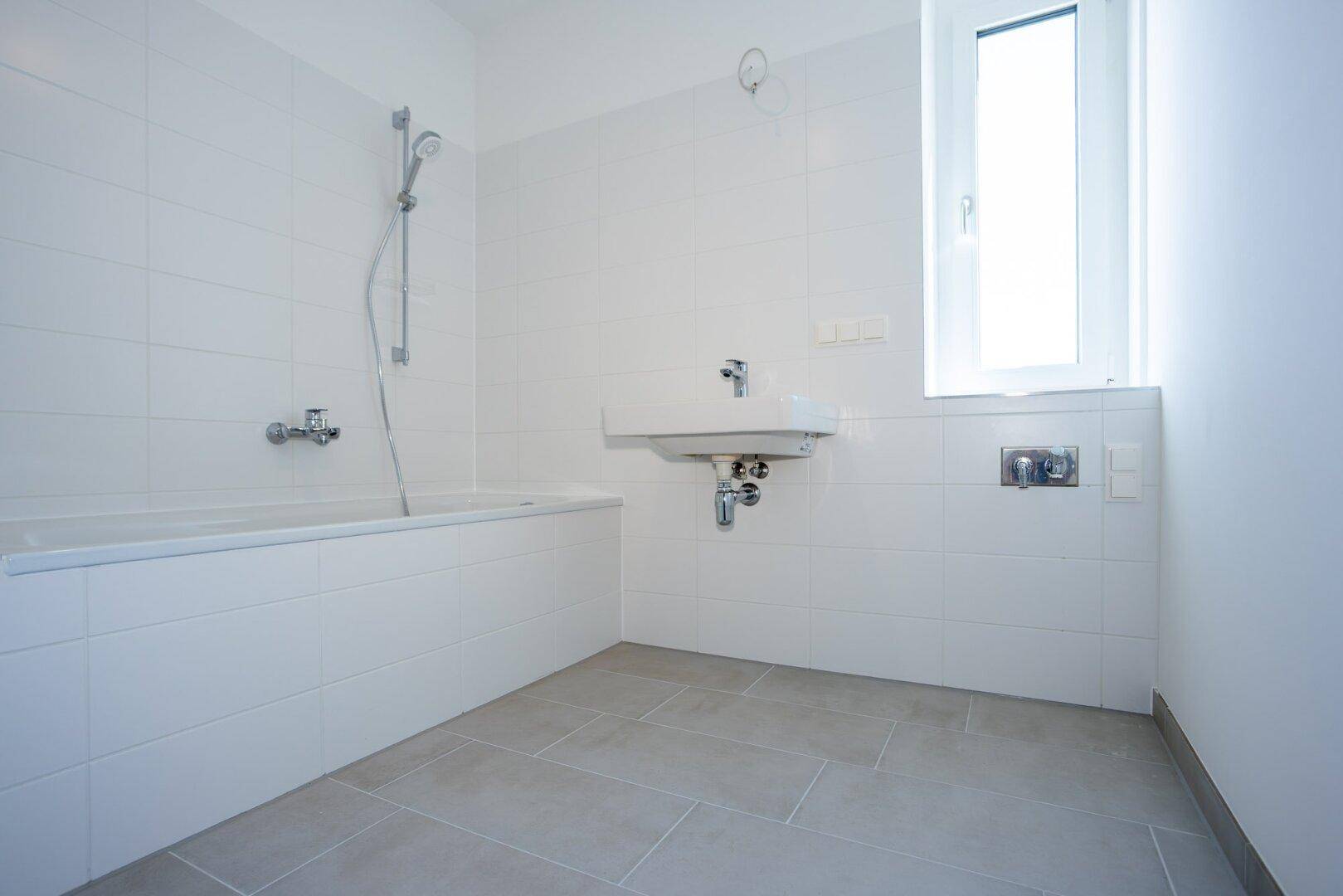 Modernes Badezimmer mit Badewanne, Waschbecken, weißen Fliesen und Fenster für Tageslicht.