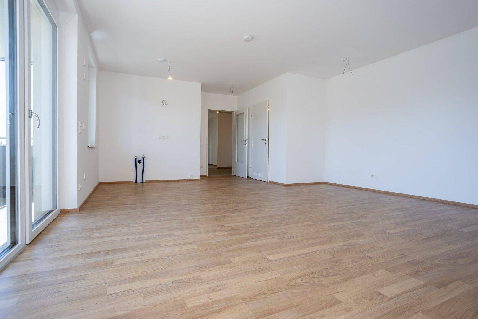 Großzügiger, heller Raum mit Laminatboden, großen Fenstern und Balkonzugang.