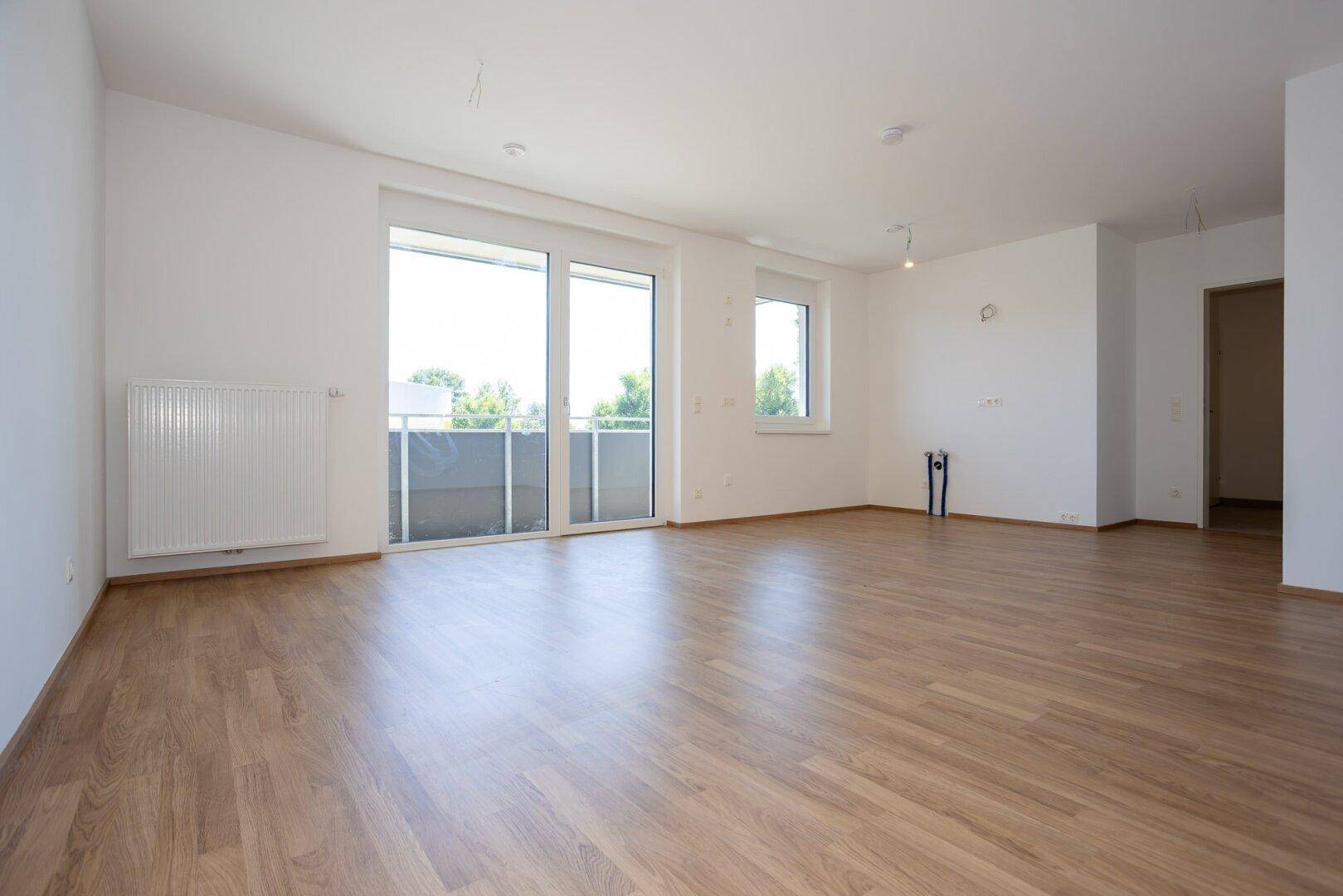 Geräumiges, leeres Zimmer mit großen Fenstern, Balkonzugang und hellem Laminatboden.