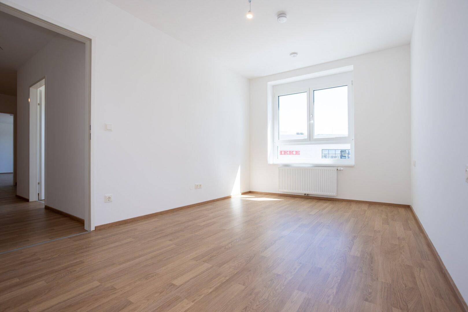 Unmöbliertes Schlafzimmer mit Laminatboden, Fenster und offenem Durchgang zu einem anderen Bereich.