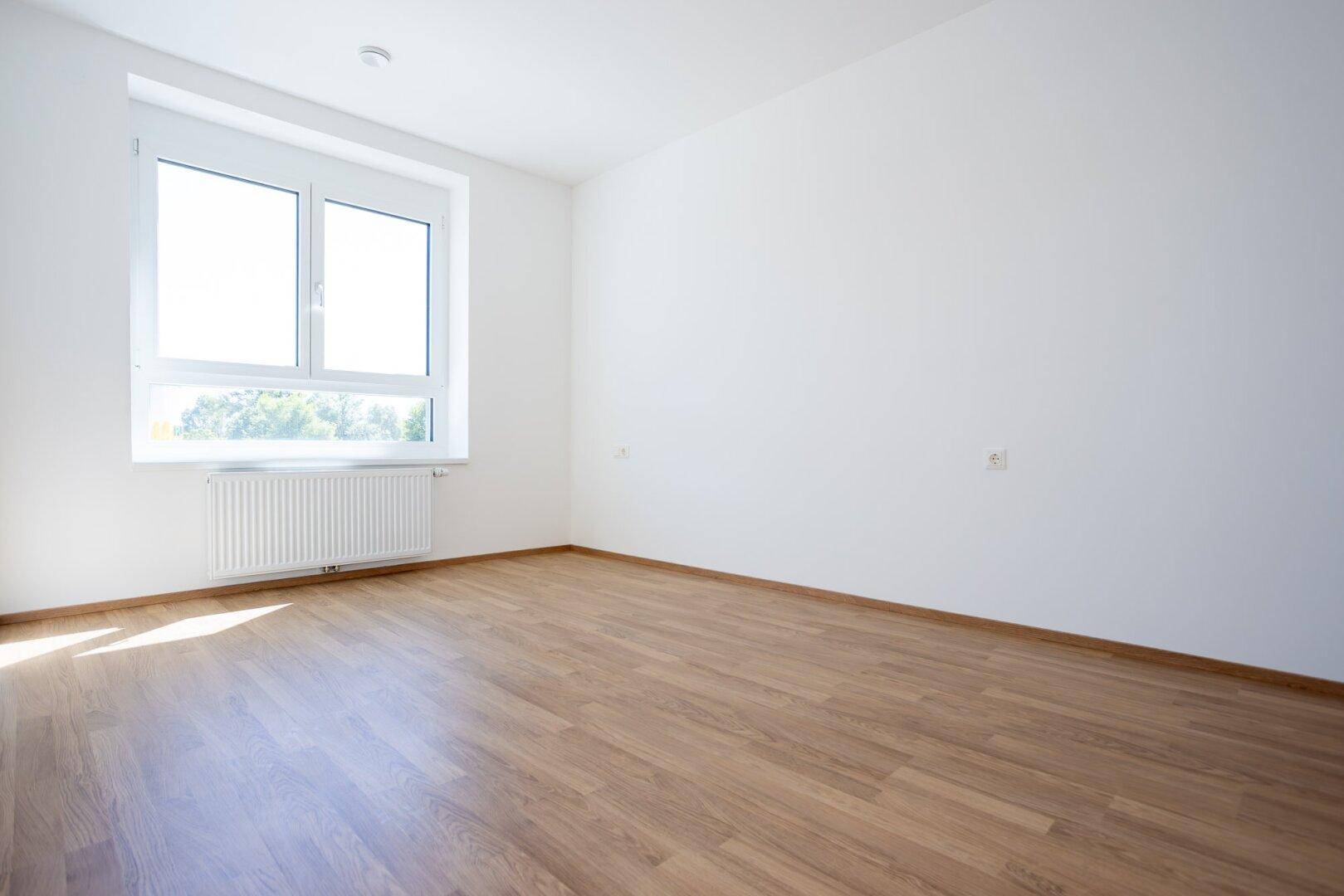 Helles Schlafzimmer mit Laminatboden und einem großen Fenster, das viel Tageslicht hereinlässt.