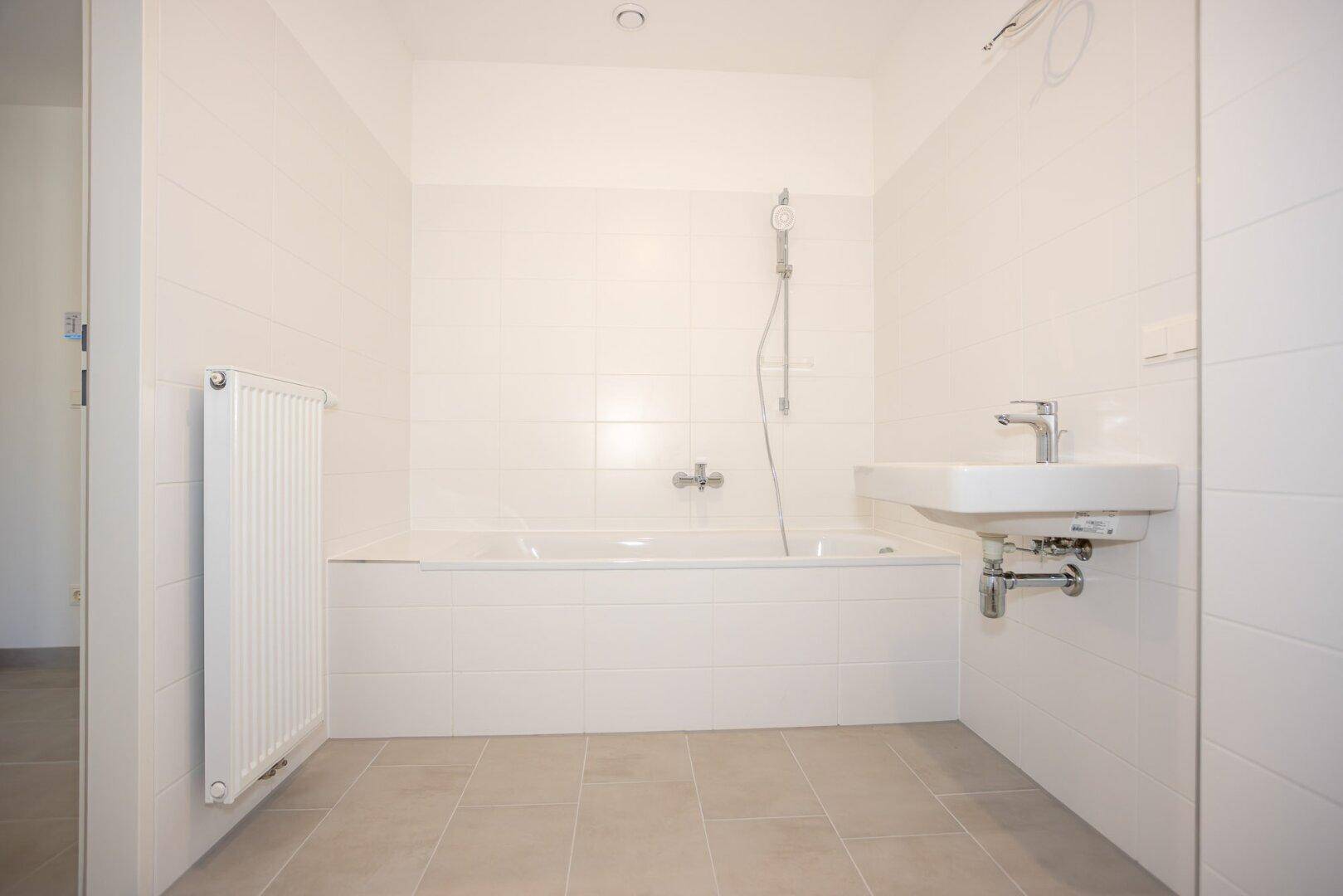 Funktionales Badezimmer mit Badewanne, Waschbecken und Heizkörper, alles in hellen Tönen gehalten.