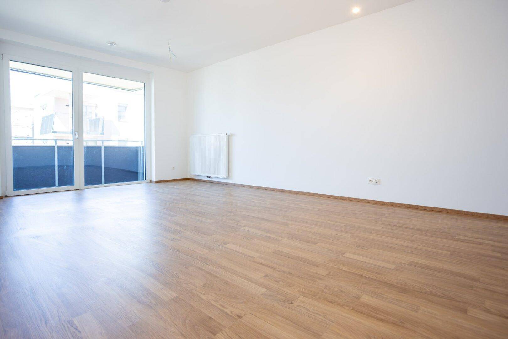 Geräumiges, unmöbliertes Zimmer mit hellem Laminatboden und großem Fenster zum Balkon.