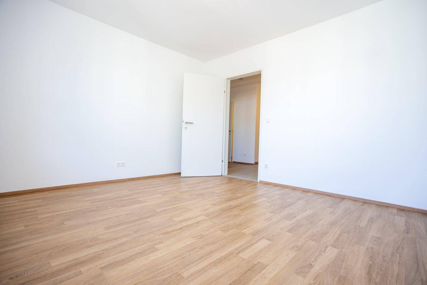 Leeres Zimmer mit hellem Laminatboden und offener Tür zu einem weiteren Raum.