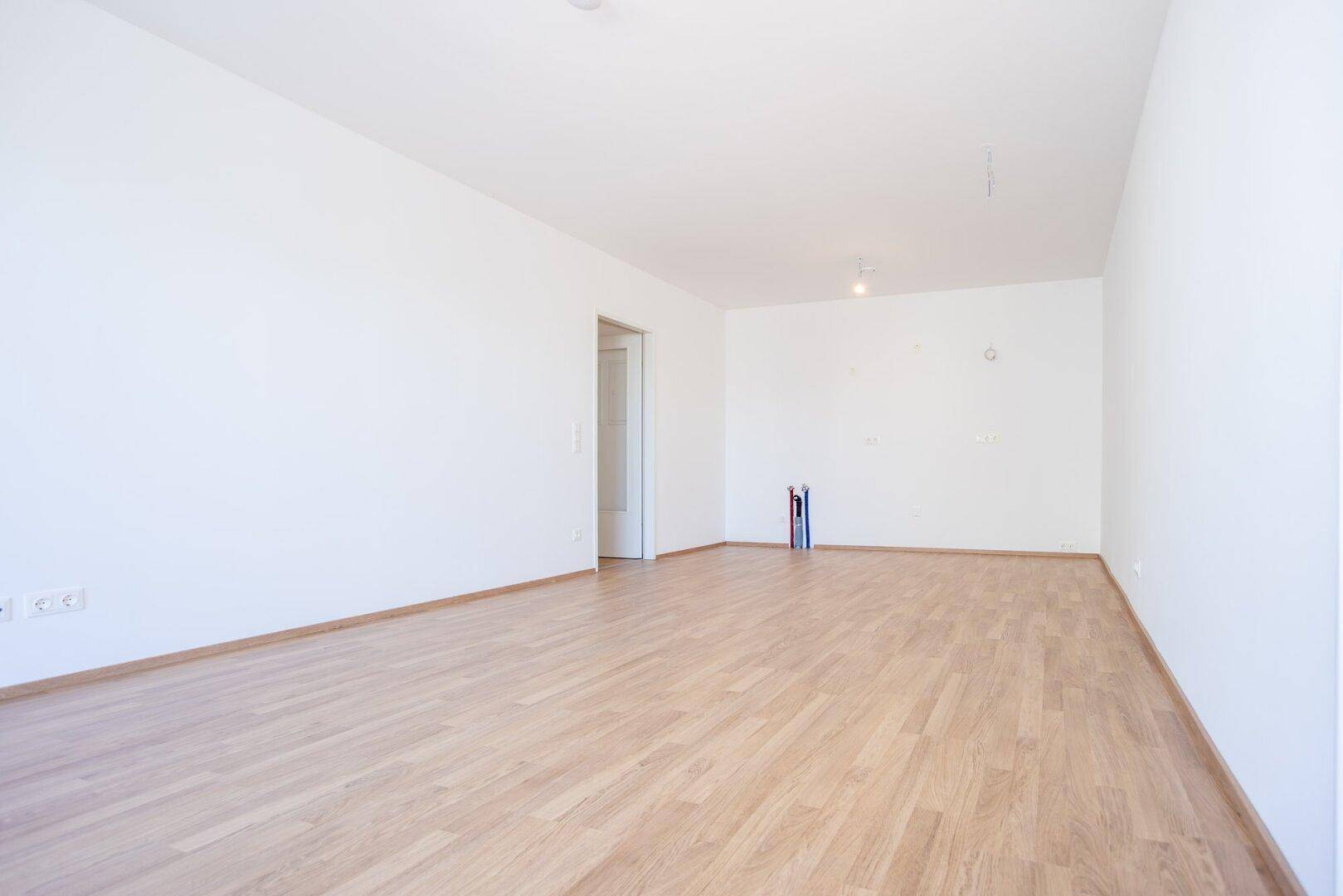 Leerer Raum mit Laminatboden und vorbereiteten Anschlüssen für eine zukünftige Küche.
