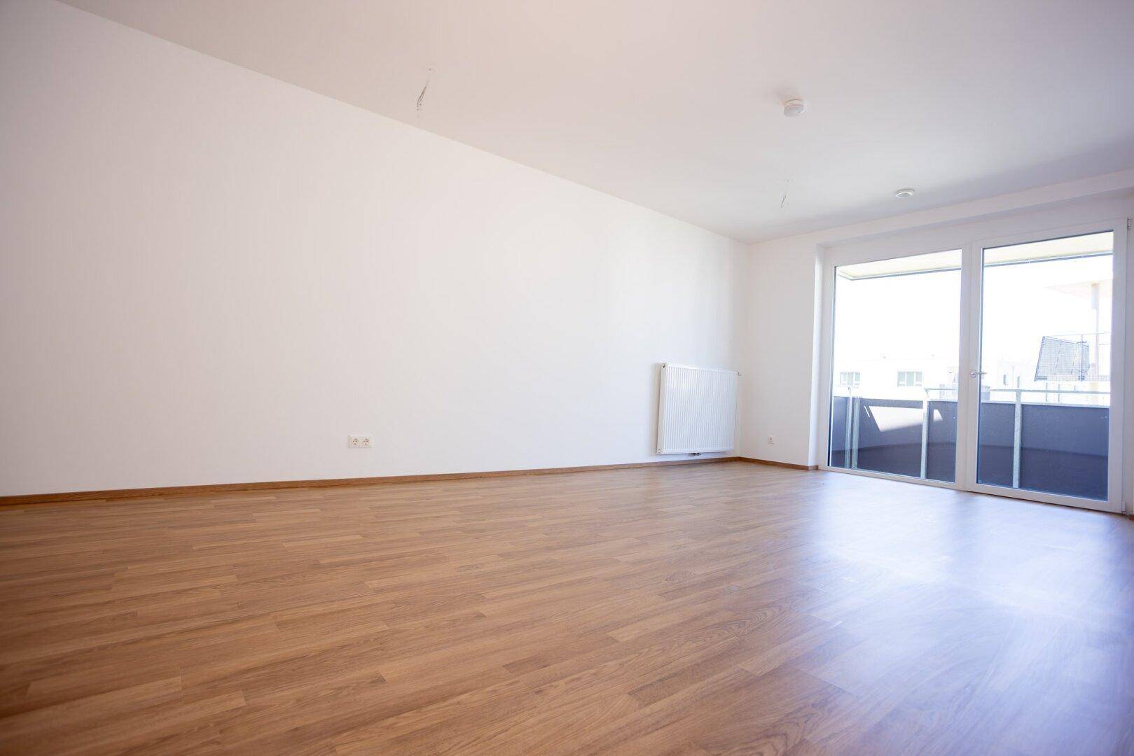 Geräumiges Wohnzimmer mit hellem Laminatboden und großen Fenstern, die viel Tageslicht hereinlassen.