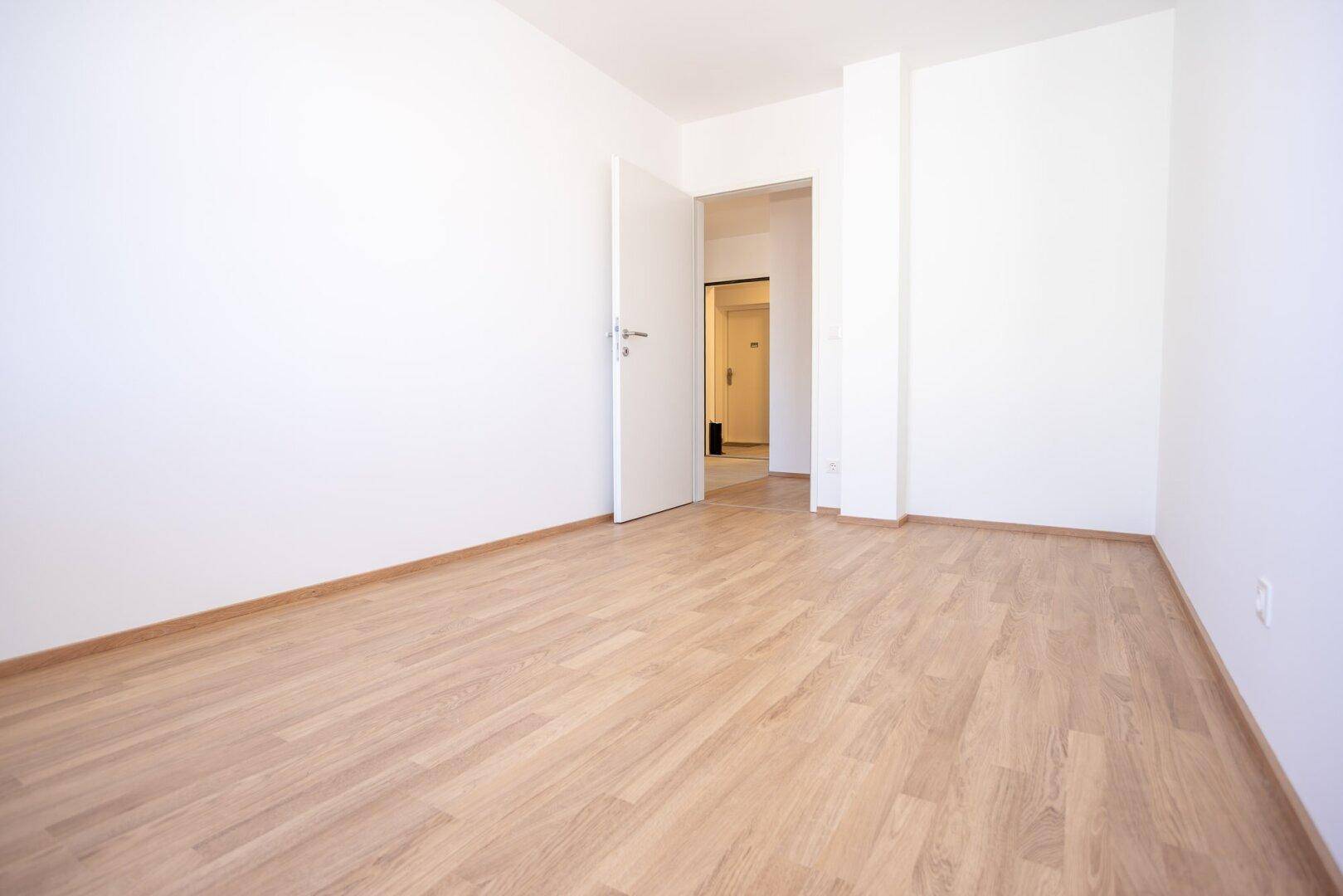 Helles Zimmer mit Laminatboden und weißen Wänden, mit Zugang zu einem Flur.