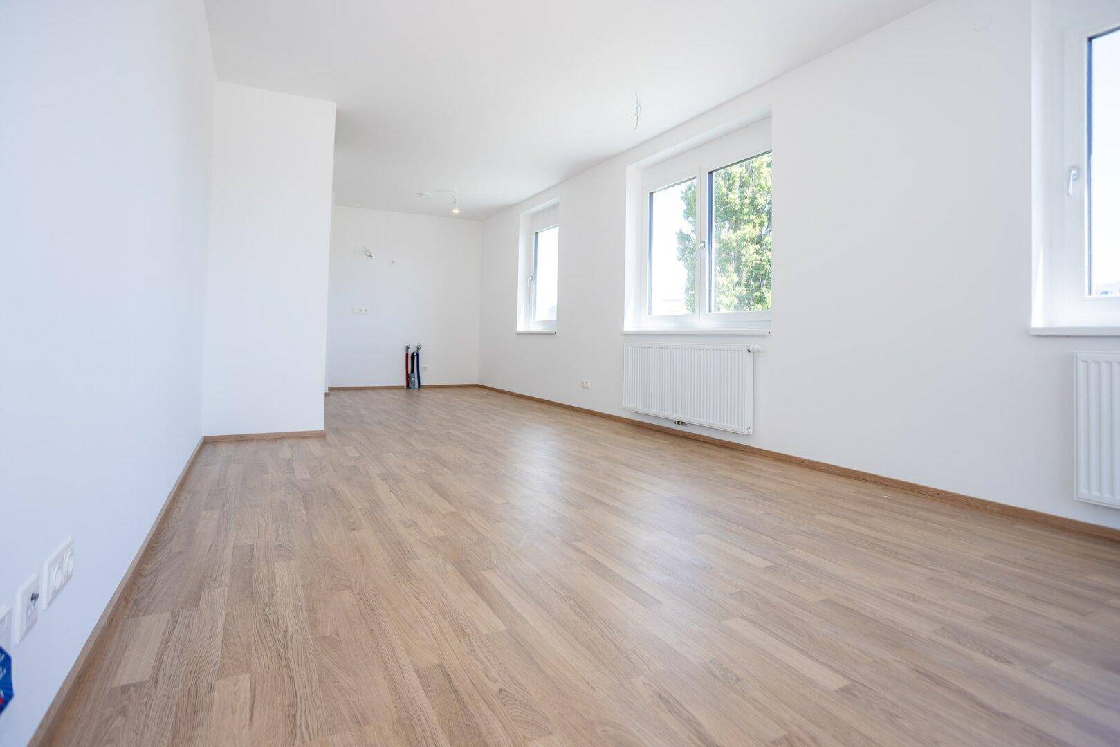 Heller, leerer Raum mit Holzboden und großen Fenstern, die viel Tageslicht hereinlassen.