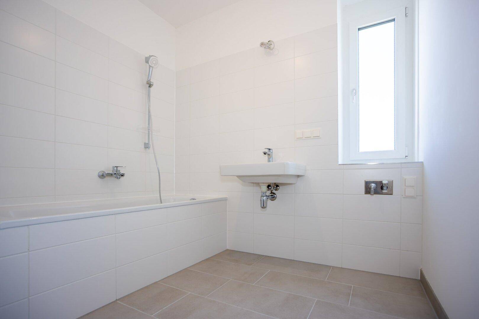 Modernes Badezimmer mit Badewanne, Einzelwaschbecken und Fenster, hell gefliest in Weiß und Grau.