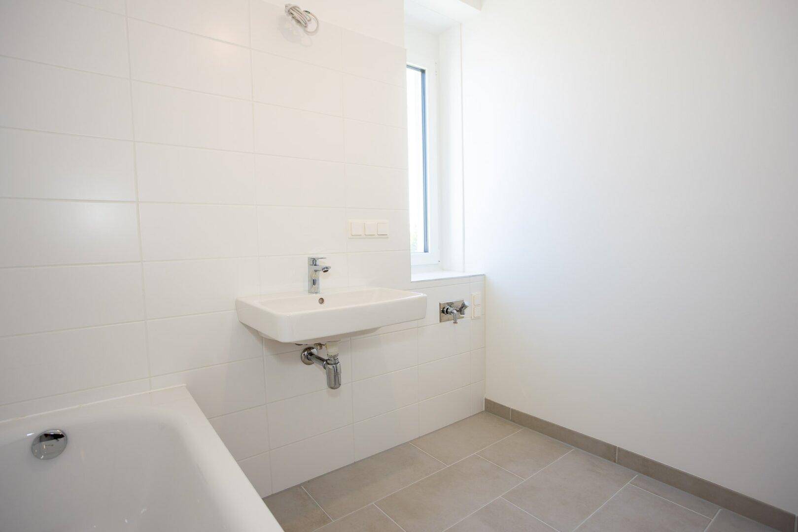 Helles Badezimmer mit Einzelwaschbecken, weißen Wandfliesen und einem Fenster für Tageslicht.