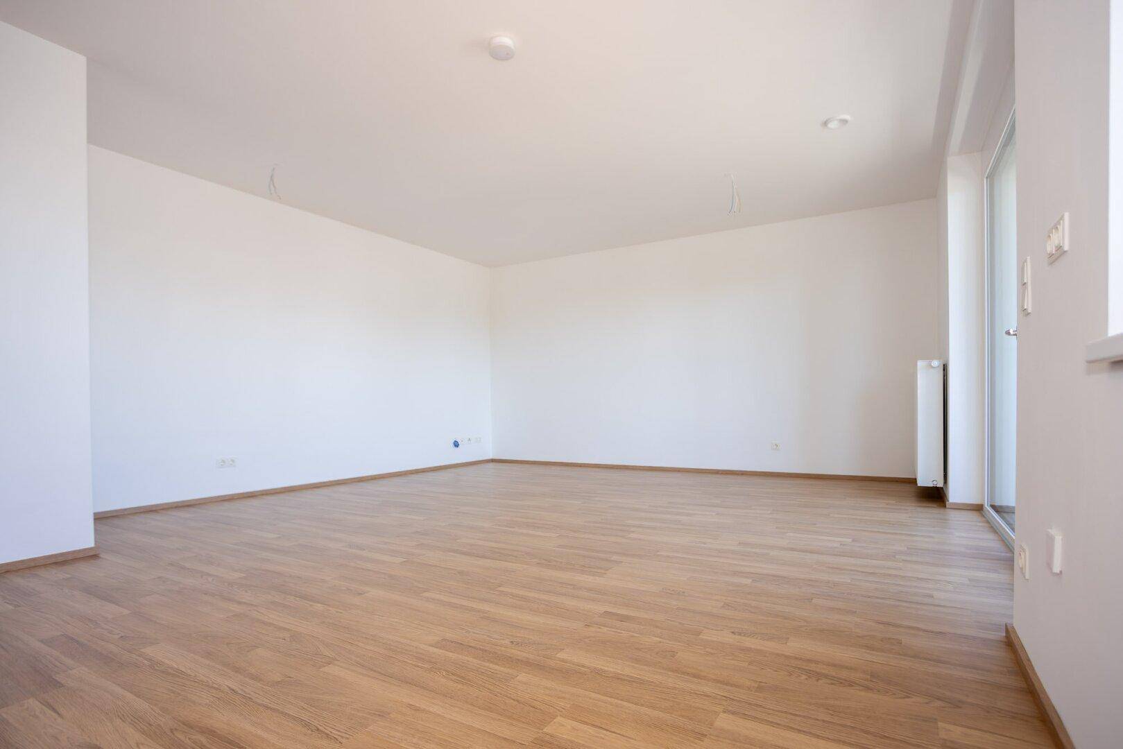 Lichtdurchfluteter Wohnraum mit hellem Laminatboden und großen Fenstern, ideal für individuelle Gestaltung.