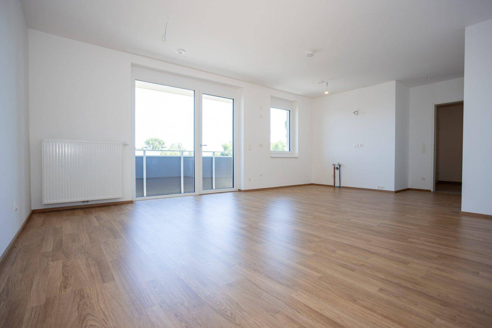Heller, leerer Wohnbereich mit Laminatboden, weißen Wänden und Zugang zu einem Balkon.