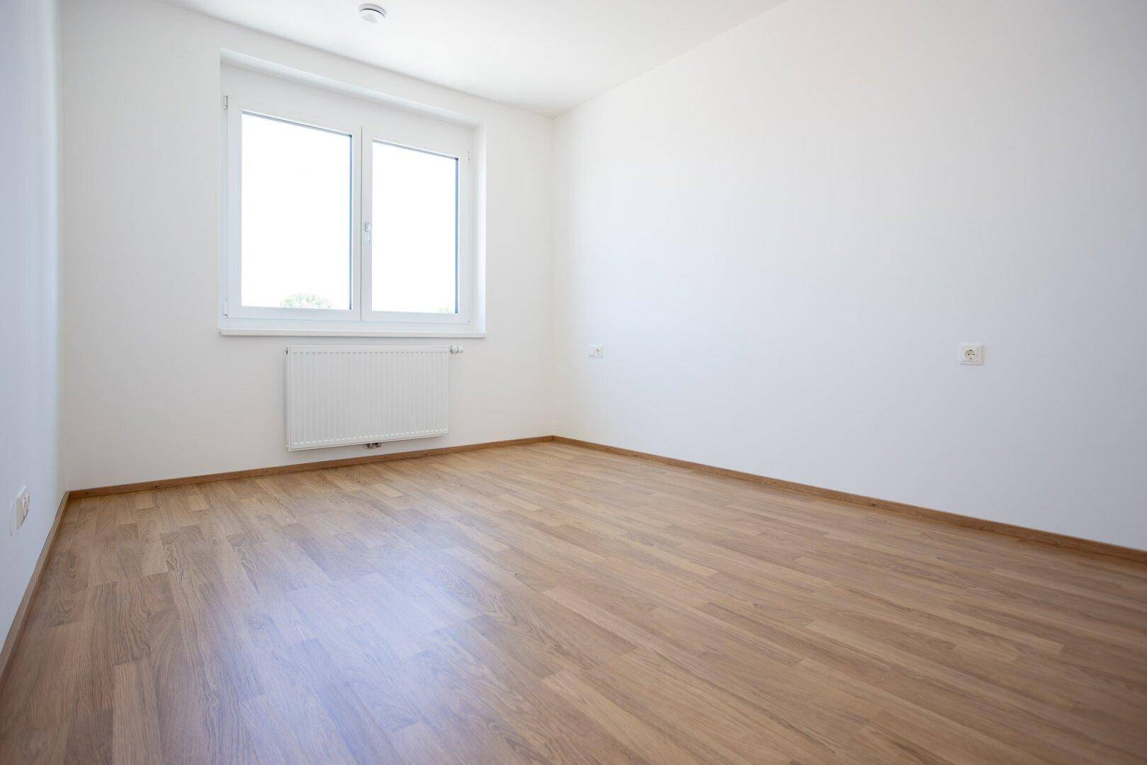 Helles Schlafzimmer mit Laminatboden und einem großen Fenster, das viel Tageslicht hereinlässt.