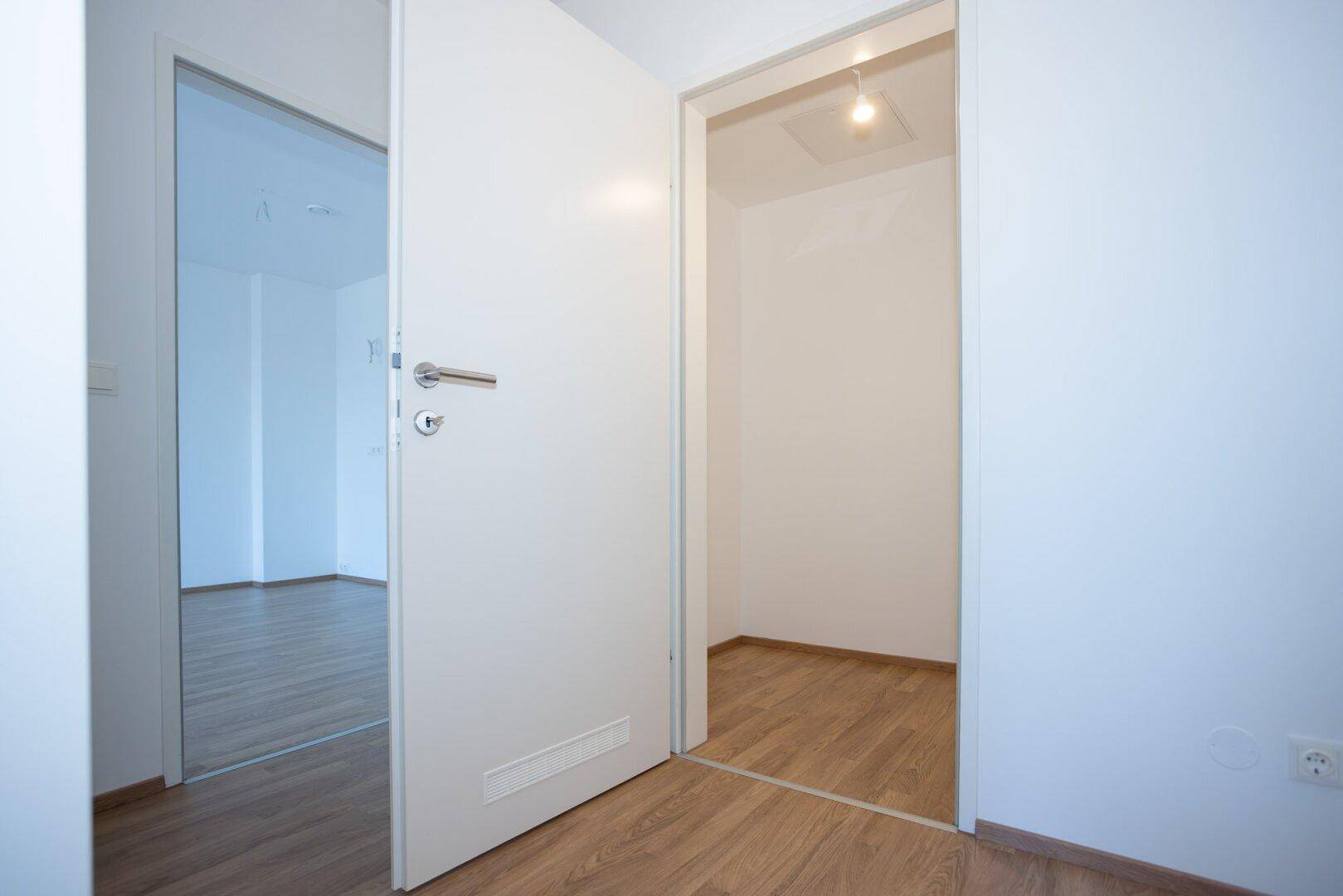 Heller Flur mit weißer Tür, die zu einem weiteren Zimmer mit Laminatboden führt.
