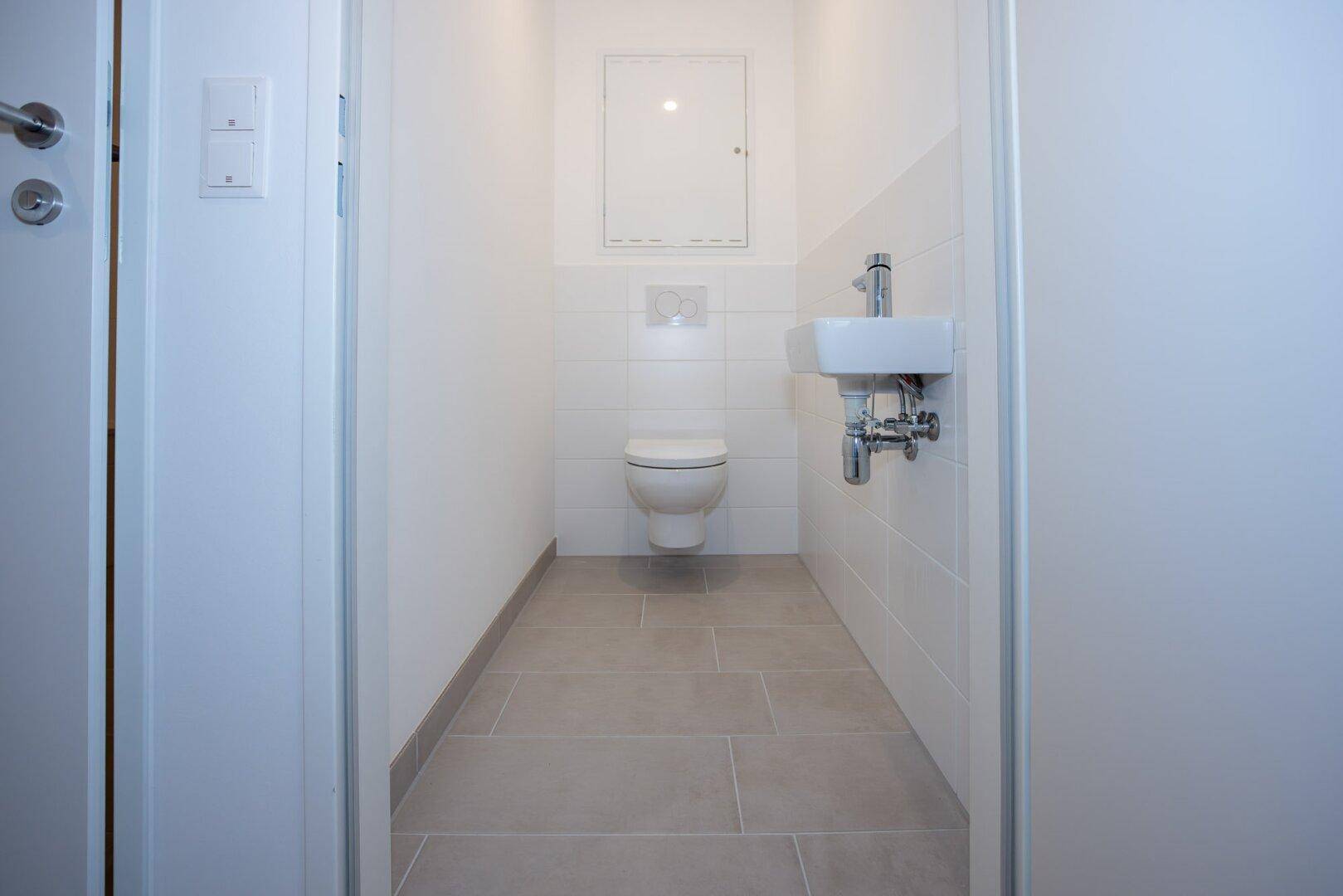 Separates Gäste-WC mit wandhängender Toilette und kleinem Waschbecken, hell gefliest.