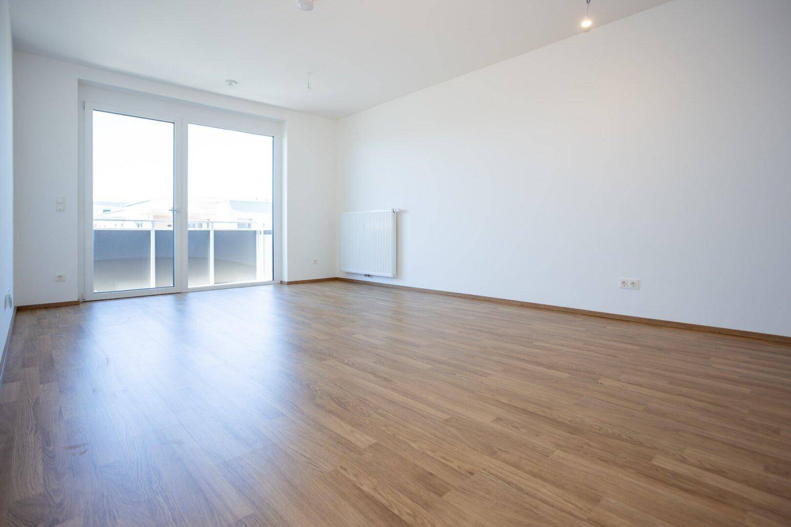 Helles Wohnzimmer mit Laminatboden und großen Fenstern, die viel Tageslicht hereinlassen.