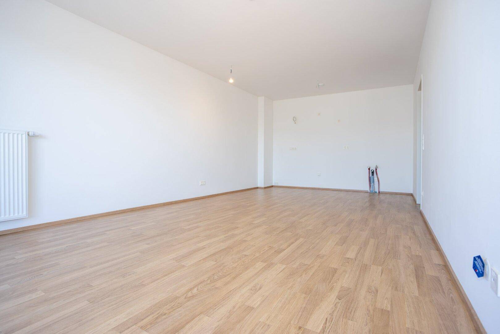 Heller, unmöblierter Raum mit Laminatboden und Heizkörper, ideal für ein Wohnzimmer.