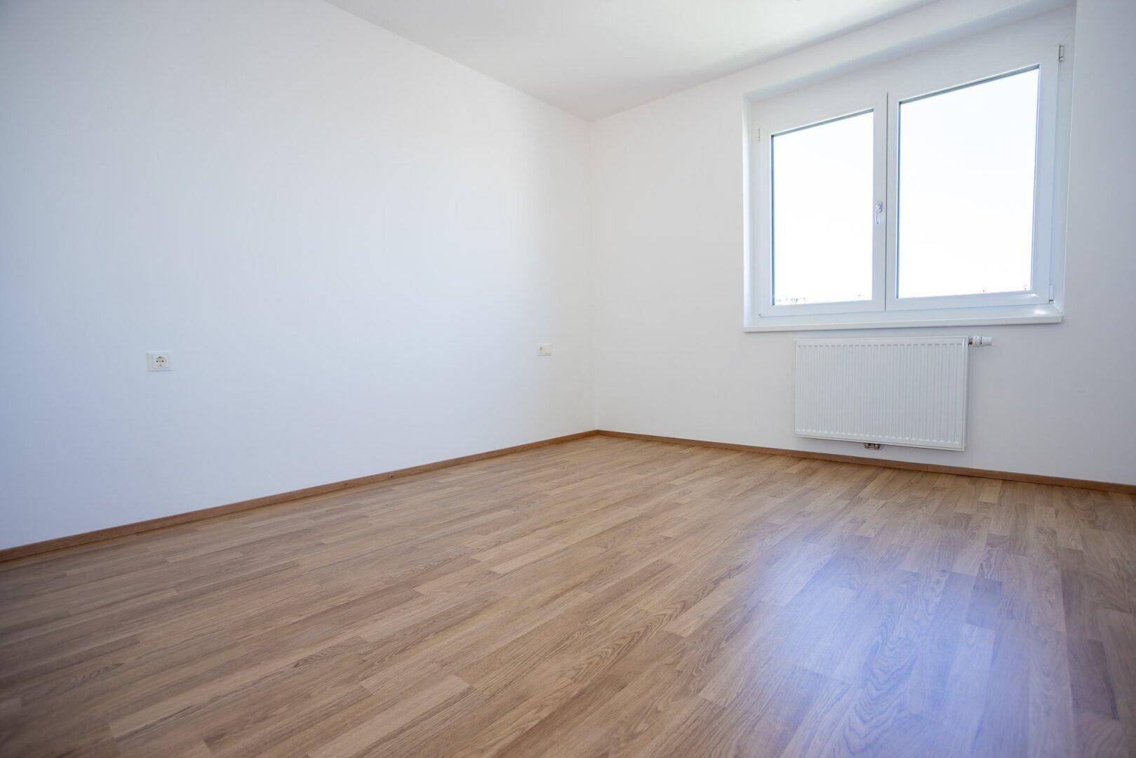 Helles Schlafzimmer mit Laminatboden und großem Fenster, das viel Tageslicht bietet.