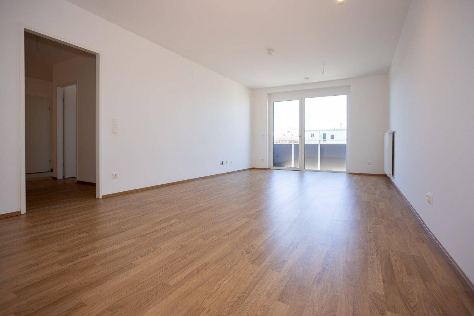 Geräumiges, leeres Wohnzimmer mit hellem Laminatboden und Zugang zum Balkon.