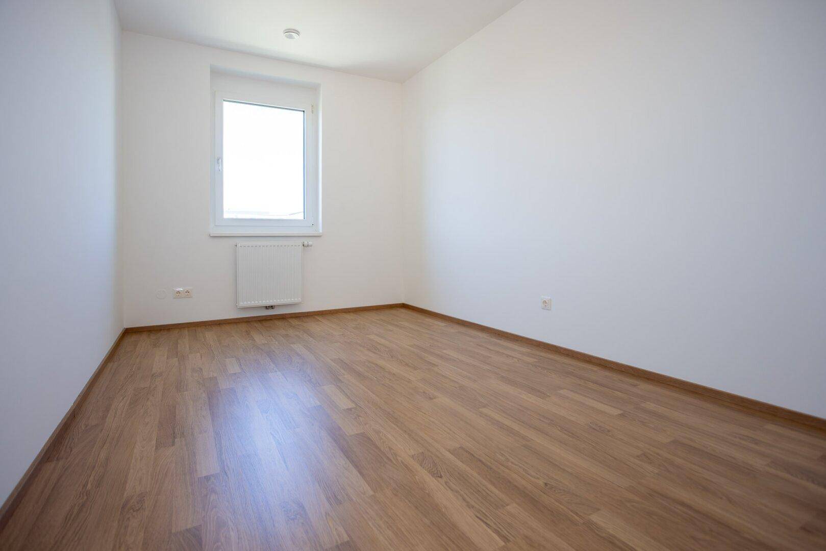 Helles Zimmer mit Laminatboden und einem einzelnen Fenster, ideal als Kinder- oder Arbeitszimmer.