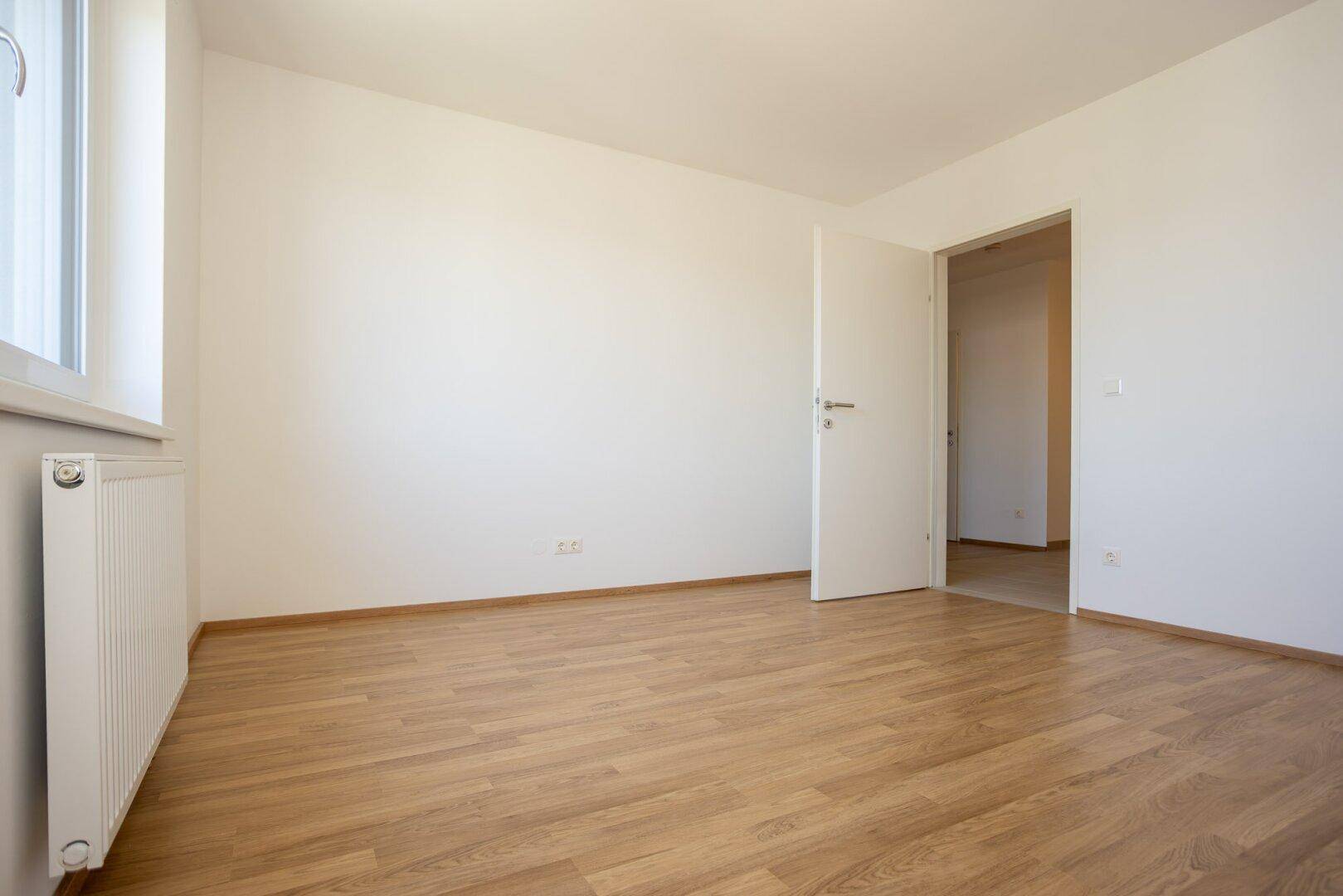 Leeres Schlafzimmer mit Laminatboden, Fenster und offener Tür zu einem weiteren Raum.