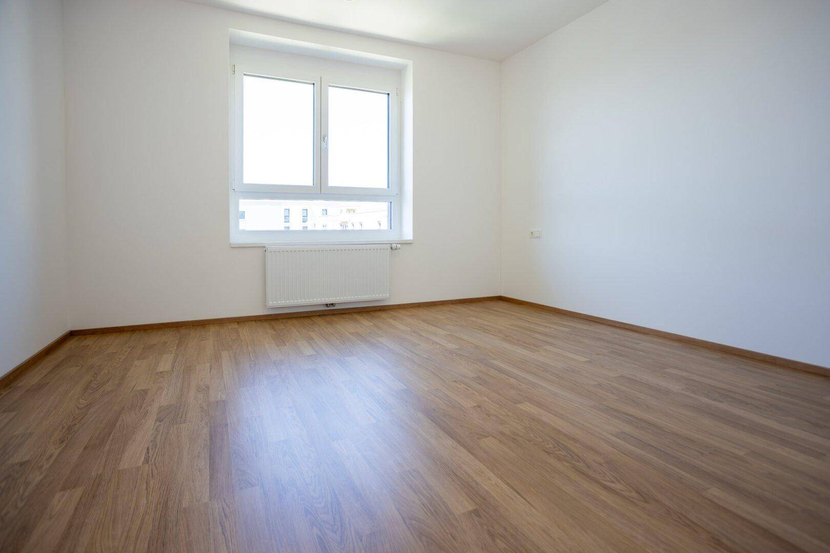 Leeres Schlafzimmer mit Laminatboden und großem Fenster, das viel Tageslicht hereinlässt.