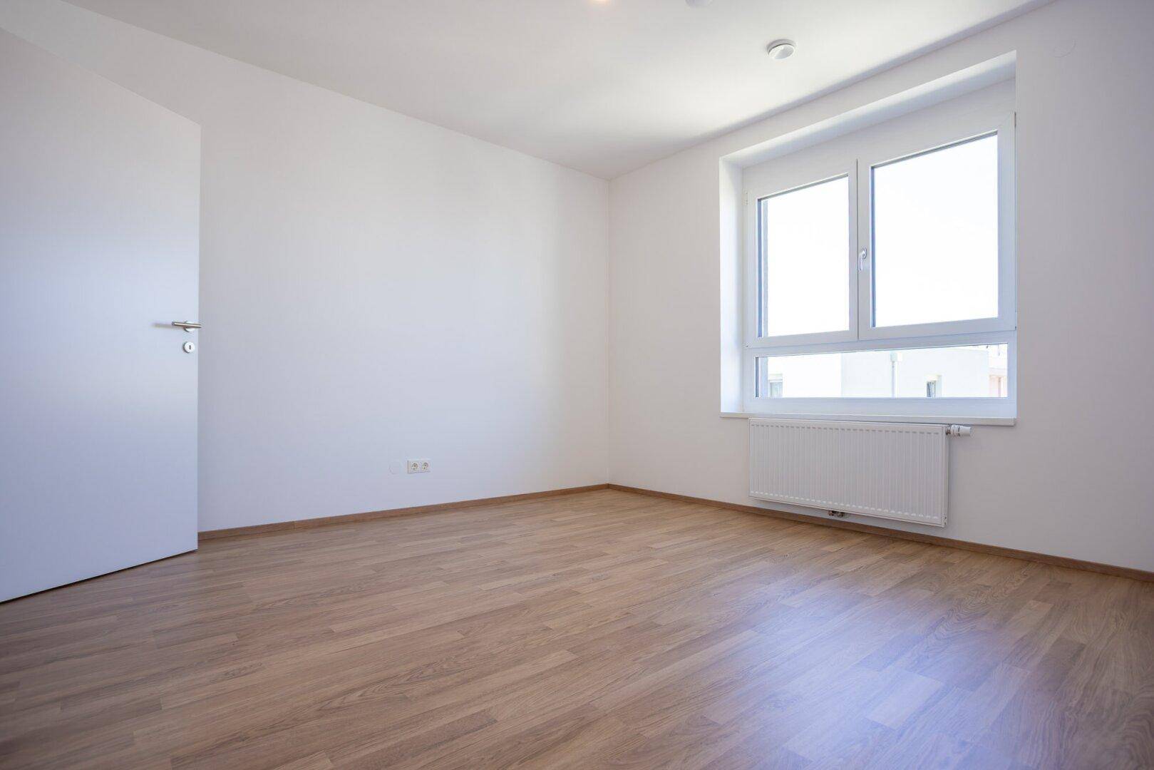 Helles Schlafzimmer mit Laminatboden und Fenster, das einen Blick nach draußen bietet.