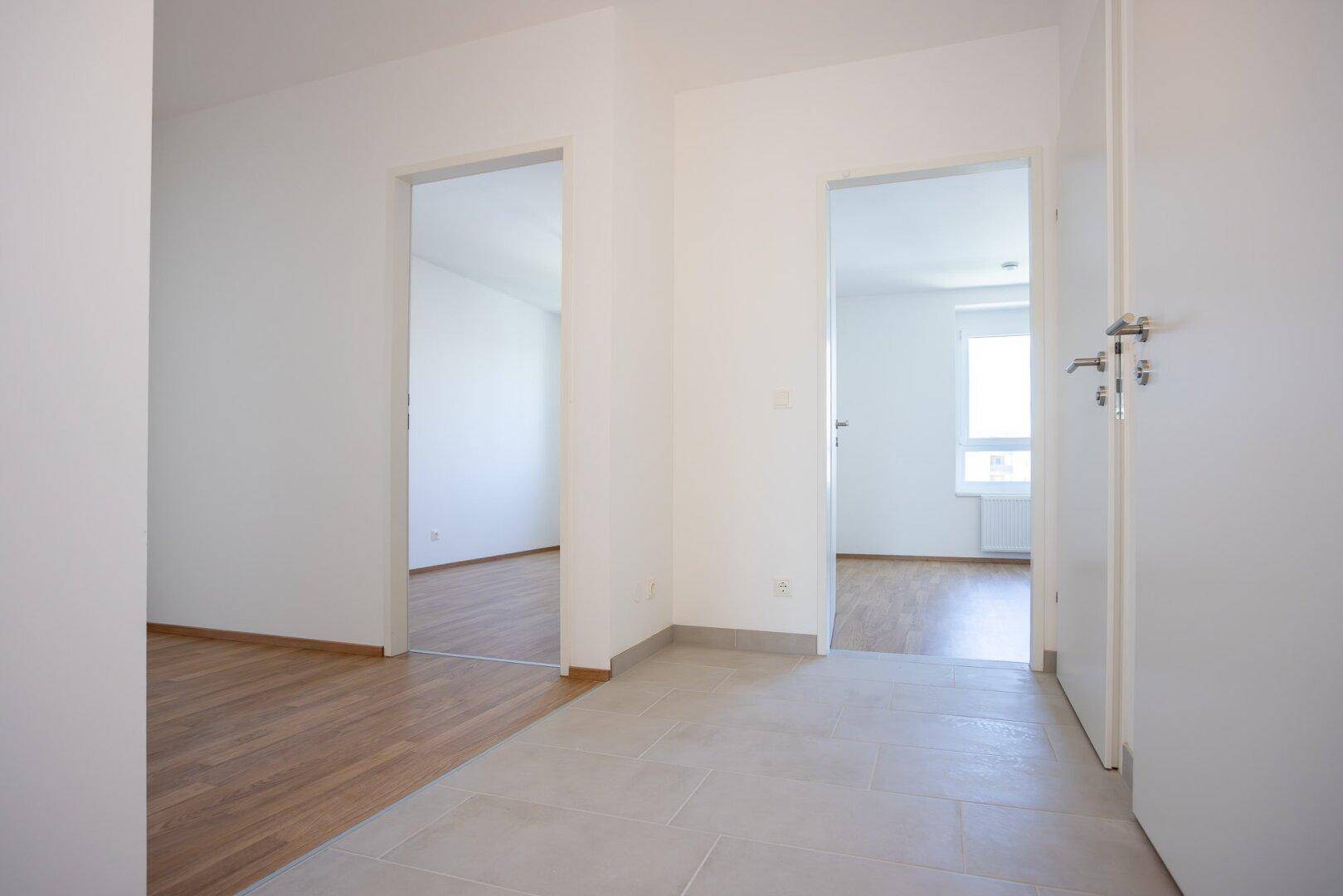 Flur mit Fliesenboden, der zu zwei hellen Räumen mit Laminatboden führt.