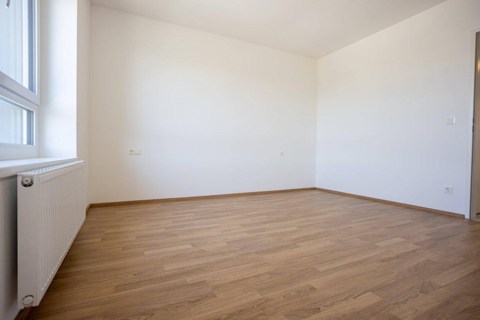Helles Schlafzimmer mit Laminatboden, weißen Wänden und einem Fenster mit Heizkörper.