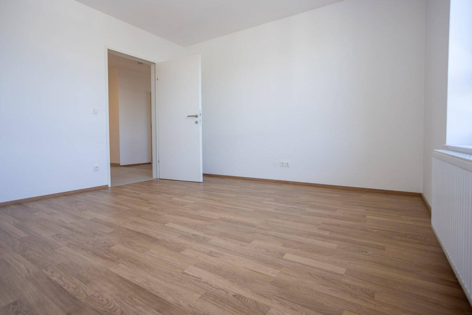 Geräumiges Schlafzimmer mit Laminatboden und weißen Wänden, mit offener Tür zu einem weiteren Raum.