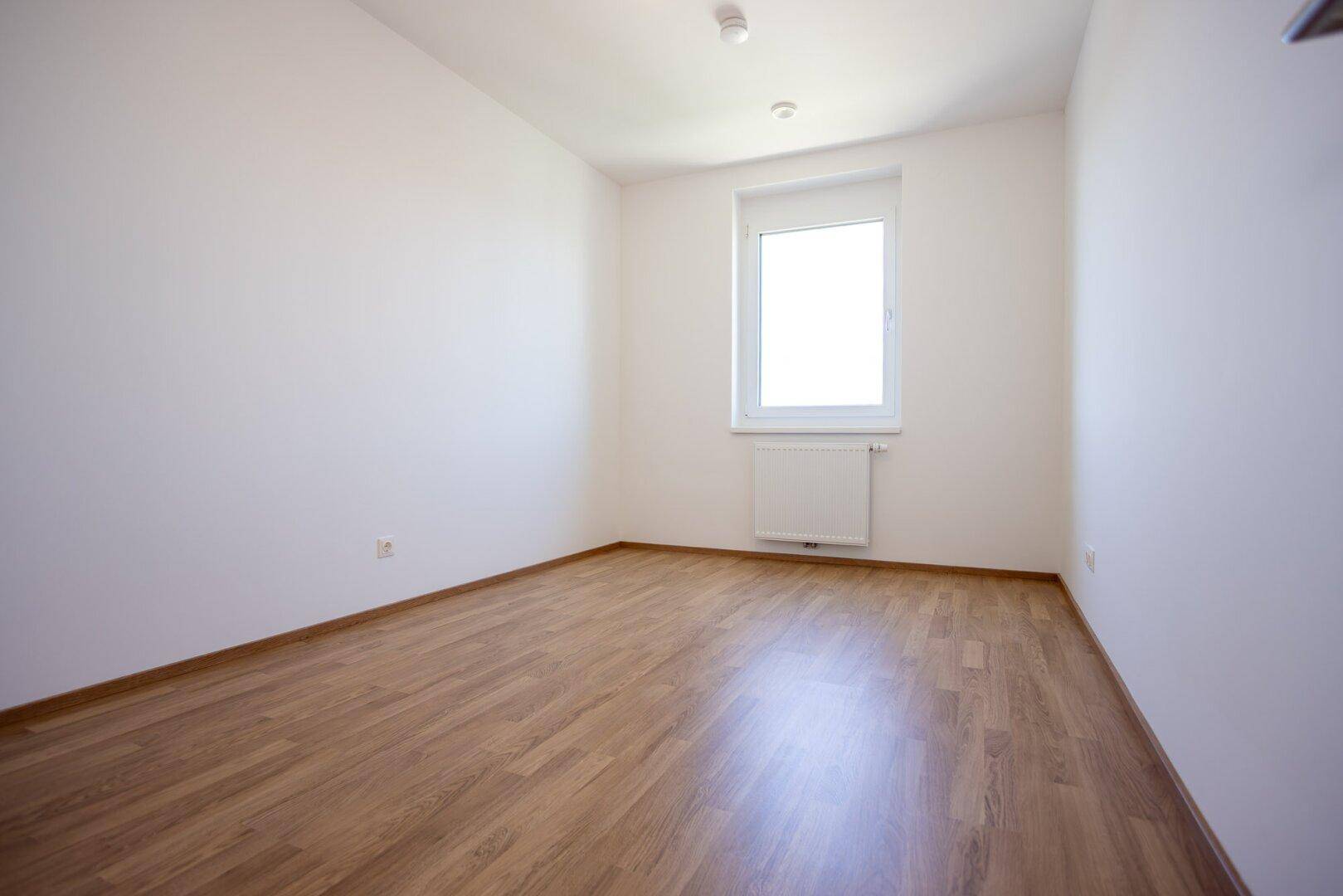 Helles Schlafzimmer mit Laminatboden, weißen Wänden und einem Fenster mit Heizkörper.