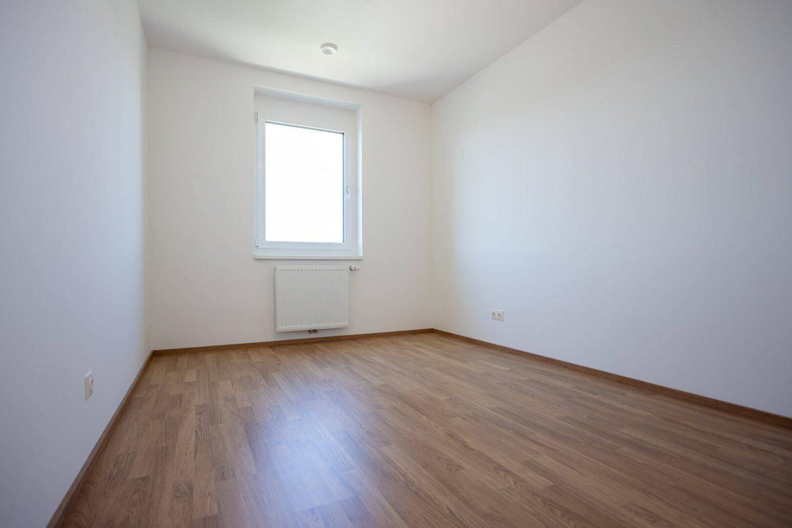Helles Schlafzimmer mit Laminatboden, weißen Wänden und einem Fenster mit Heizkörper.
