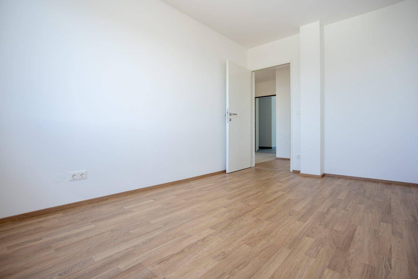 Geräumiges Schlafzimmer mit Laminatboden und weißen Wänden, mit offener Tür zu einem weiteren Raum.